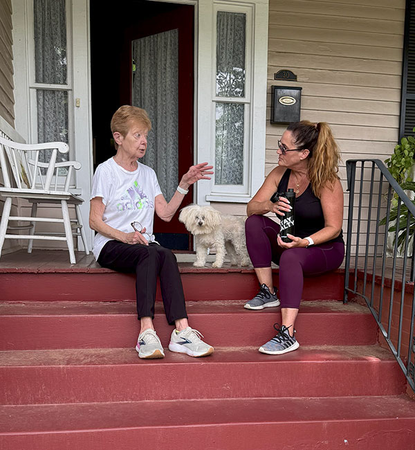 Nora sitting on the red steps of a porch listening to a woman sitting beside her talk with a white poodle between them